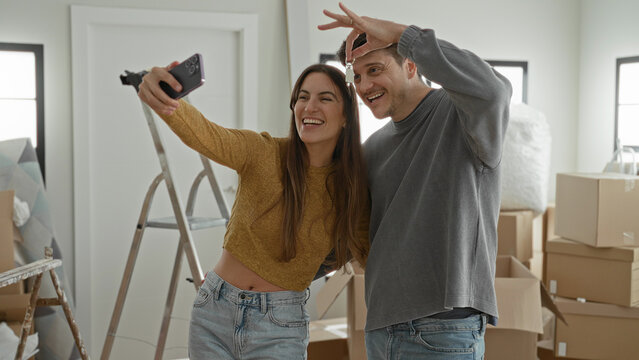 Young woman and man smile and hold smartphone for selfie amid packed boxes and ladder in a building; joy new home together.
