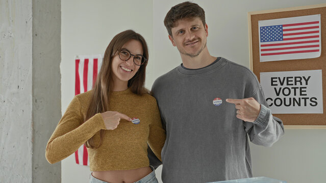 Young woman points finger to voted sticker on chest beside partner at ballot box in polling station building; civic engagement pride.