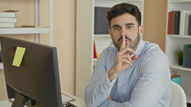 Man at computer with finger to lips performing shushing gesture in office building; quiet concentration.