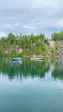 Three inflatable boats in calm lake water surrounded by lush green trees and rocky cliffs under a cloudy sky in a serene natural landscape. Slow motion vertical footage. Nature concept. Russia
