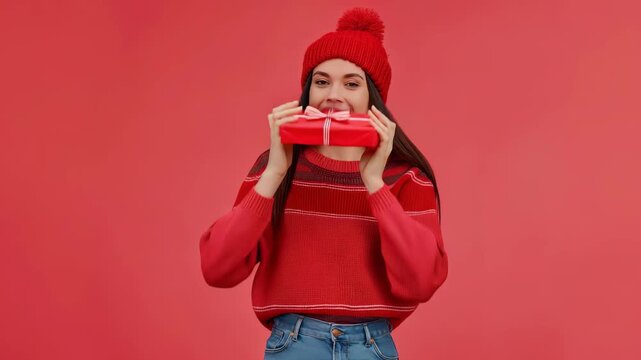 An elated woman in a red hat and matching sweater excitedly holds a red gift box over her eyes. Set against a vibrant red backdrop, the image conveys a sense of joy and festivity.