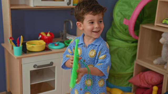 Boy blows a green plastic toy trumpet while smiling in a playroom by a play kitchen and shelves; playful joy.