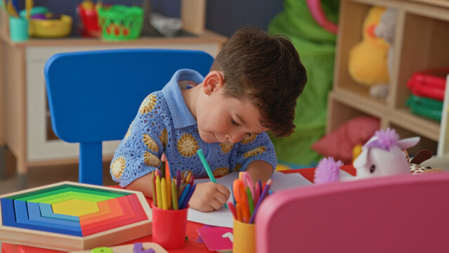 Little boy intently drawing with colored pencils on paper at a small craft table in preschool building; creativity concentration.