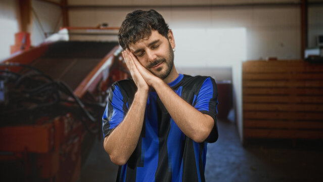 Man with beard wearing blue football shirt presses hands to cheek in a sleeping gesture inside industrial building; sleepy rest.