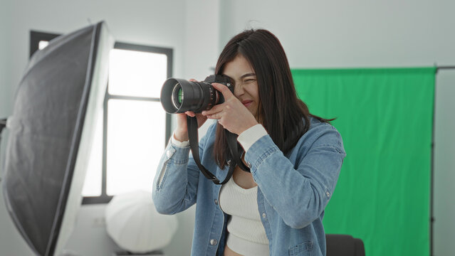 Young woman holds camera and gestures with her hand while smiling in a studio green screen setup; confidence creativity.