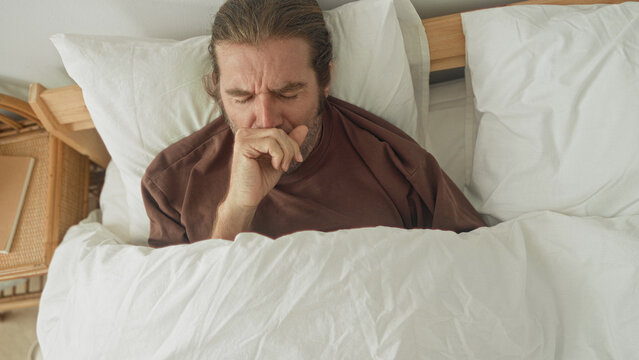 Man covering mouth yawning in bed under white duvet with pillow on bedside table inside a building; fatigue morning rest contemplation.