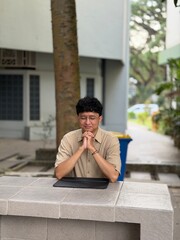 Obraz premium A young man with curly black hair sits outdoors with hands clasped in prayer, surrounded.