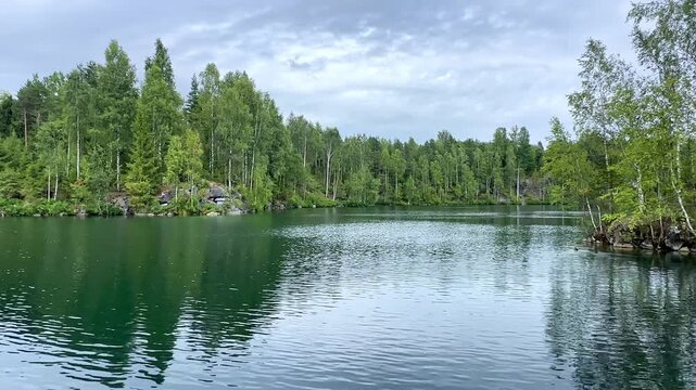 Serene lake surrounded by lush green trees and rocky shoreline under a cloudy sky in a tranquil natural setting. Ruskeala Mountain Park in Karelia, Russia.