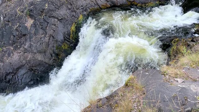 Water flows rapidly over dark rocks in a natural environment, with patches of grass and vegetation visible along the banks, highlighting contrast between the water and rocky surface. footage