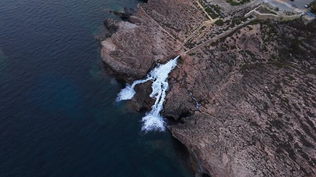 Industrial wastewater cascades from a reverse osmosis desalination plant outlet down rugged limestone cliffs into the sea, captured by drone above Ghar Lapsi, Malta.