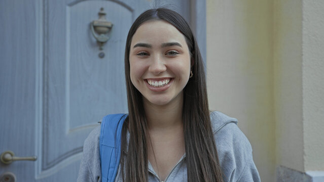 Young hispanic woman student wearing gray hoodie with blue backpack smiles at camera showing teeth and dimples at a building entrance doorway; optimism new start.
