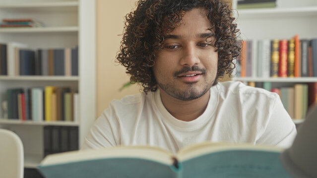 Man reads book and turns a page at a table in a library building with bookshelves behind him; concentration study.