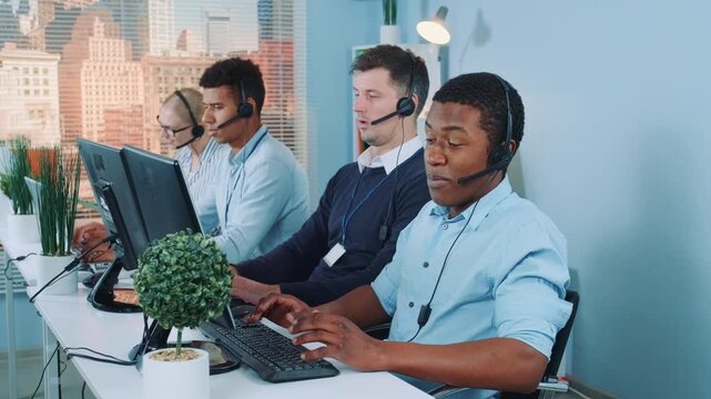 A multiracial group of five customer support agents wearing headsets working at desks in a modern office call center with cityscape views through large windows.