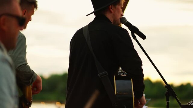Outdoor sunset performance with guitarist singing into microphone on riverside stage, accompanied by band members playing instruments during relaxed live concert