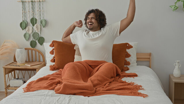 Young indian man on bed stretches arms up, yawning with orange blanket, tassel pillow and wicker bedside table in studio; morning serenity.