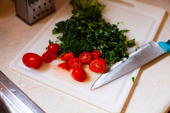 Chopped cherry tomatoes and fresh green herbs rest on a white plastic cutting board in Corfu,Greece. A stainless steel kitchen knife sits beside the prepared ingredients on the counter.
