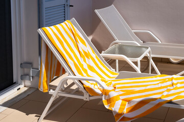 White sun loungers with yellow striped towels rest on a tiled resort balcony in Corfu,Greece. A small side table stands between the chairs under bright direct sunlight. © dsbedarieva