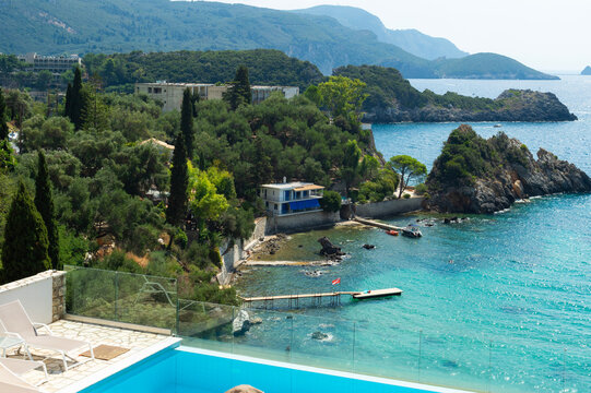 An outdoor swimming pool and lounge terrace overlook a turquoise sea bay in Corfu,Greece. Coastal houses, green mountains, and a pier border the clear blue waters.