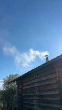 Real-time 4K video of white smoke coming out of a chimney on a rustic wooden sauna roof against a blue sky.