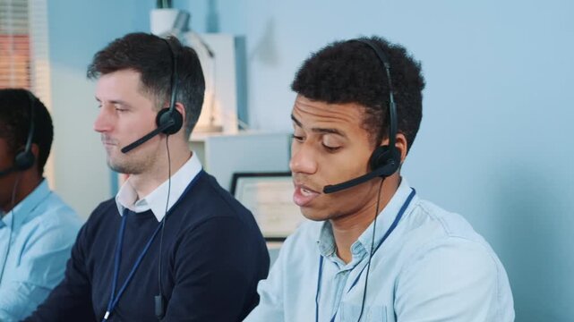 Four men wearing headsets and business casual attire focused on their tasks in a call center office with computers and simple furniture indoors.