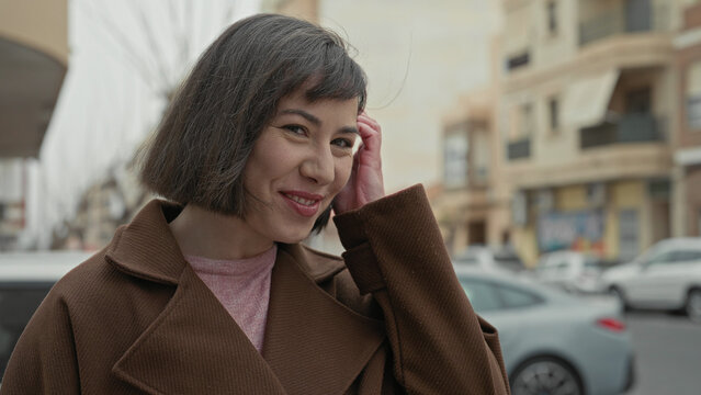 Woman smiling and tucking hair with hand while wearing a coat on street, parked cars and shops visible; gentle contentment.