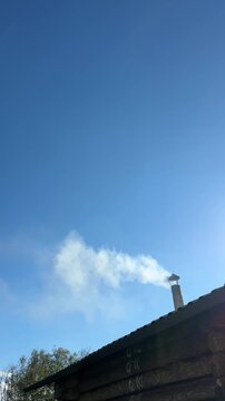 Real-time 4K video of white smoke coming out of a chimney on a rustic wooden sauna roof against a blue sky.
