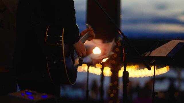 Silhouetted guitarist and singer performing live on an outdoor waterfront stage at twilight, illuminated by warm stage lights against a dramatic evening sky