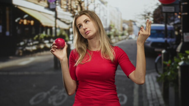 Blonde woman in a red top holding a pomegranate in her right hand while shrugging and rolling eyes on a street lined with cafes and a van; indifference doubt.