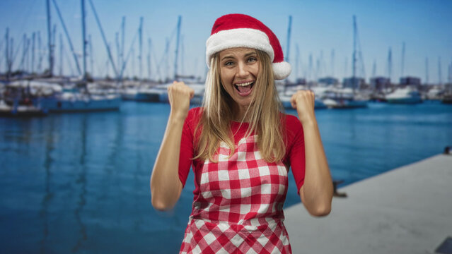 Woman wearing santa hat and checkered apron pumps fist on marina dock beside sailboats while smiling broadly; holiday joy.