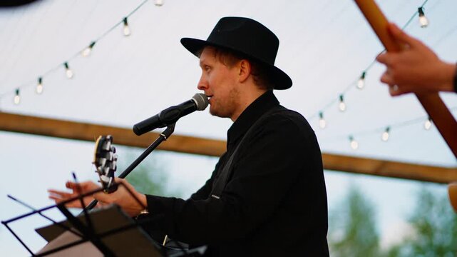 Smiling guitarist in black hat performing live on outdoor stage under hanging string lights during warm evening acoustic concert with relaxed audience