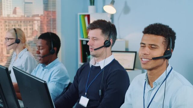 A group of five customer service agents wearing headsets working at their desks in a modern office with city views through large windows during daytime.