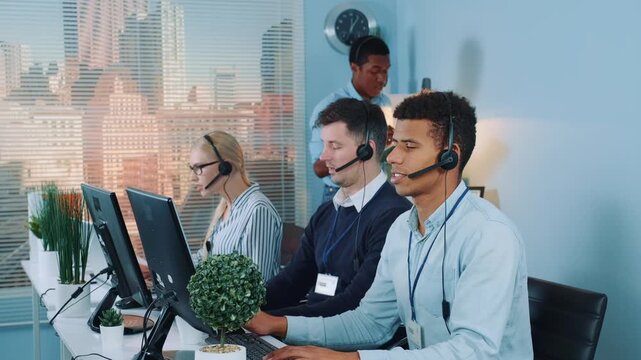 Six call center employees wearing headsets work at desks in a bright office with large windows overlooking a city skyline during daytime.