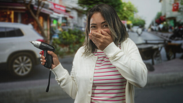 Hispanic middle aged woman holding a cordless screwdriver, hand covering mouth gesture, looking startled on a busy street with storefronts; surprise concern.