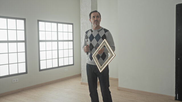 Man holds hammer and diamond picture frame in hand while looking up at a wall in an empty building; contemplation home repair.