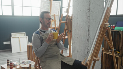 Man painter in apron sits at easel with palette, holding yellow coffee cup, hand on chin in studio  contemplative calm. © Krakenimages.com