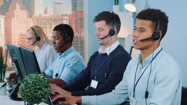 Five multiracial call center agents wearing headsets work at desks with computers in a bright office overlooking a dense urban cityscape during daytime.