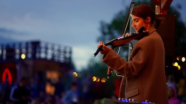 Outdoor evening musical performance featuring a focused violinist and energetic singer on stage under warm festival lights with a relaxed crowd in the background
