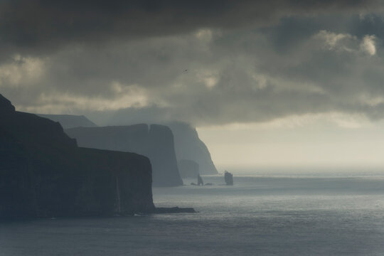 Rugged coastal cliffs and distant sea stacks along an ocean shoreline under dark overcast storm clouds with hazy atmospheric mist.