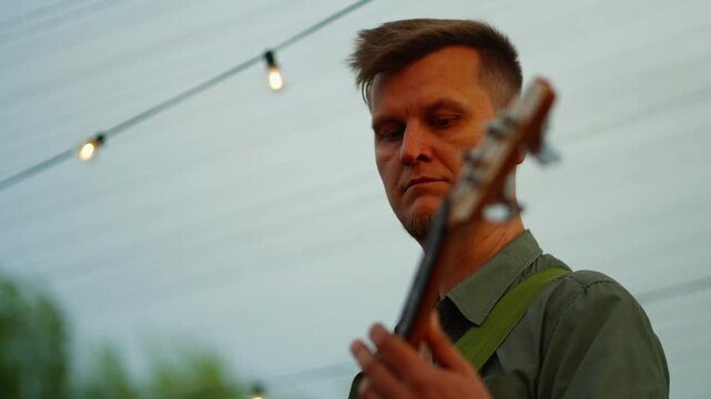 Thoughtful musician playing electric bass guitar on outdoor stage at dusk with warm string lights glowing softly and creating a relaxed, intimate concert atmosphere