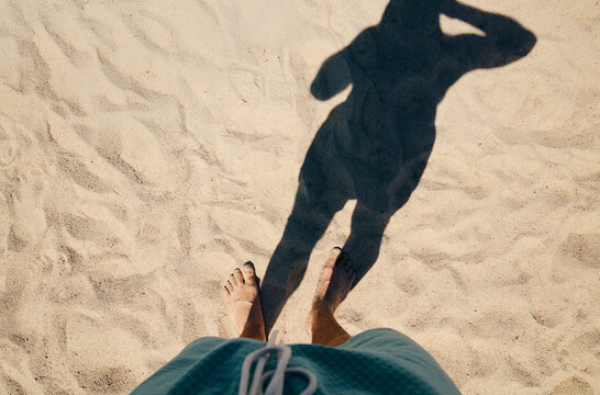 Low section POV of a man standing barefoot on sunny sandy beach casting a shadow