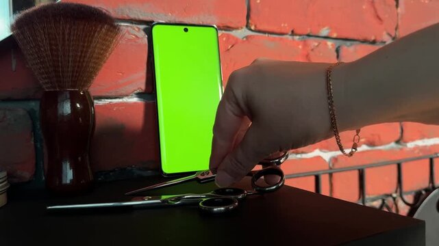 Horizontal close-up video of a hand placing professional barber scissors on a dark salon counter beside another pair, a smartphone with green chroma key screen and large neck duster by a brick wall