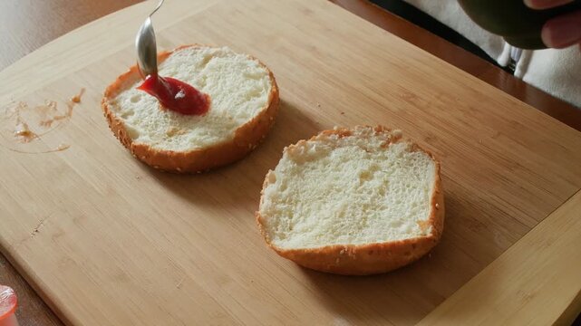 Person spreads red jam ketchup onto fresh bread slice using a knife on wooden board, demonstrating food preparation in domestic kitchen setting with natural lighting.