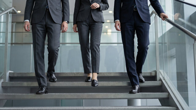 ガラスの手すりがあるオフィスの階段を下りる三人の会社員と、灰色や紺のスーツに合わせた革靴