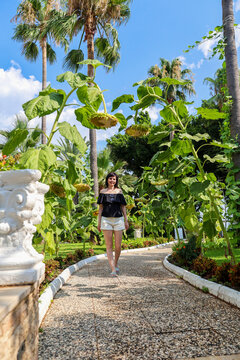 Portrait of an adult woman in off-the-shoulder clothing, a sun-drenched alley among blooming yellow sunflowers and tall palm trees.