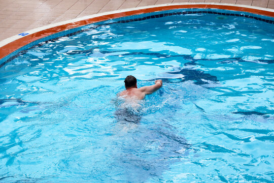 Happy man swimming and relaxing in his own small private backyard swimming pool near a house on a sunny warm summer day.