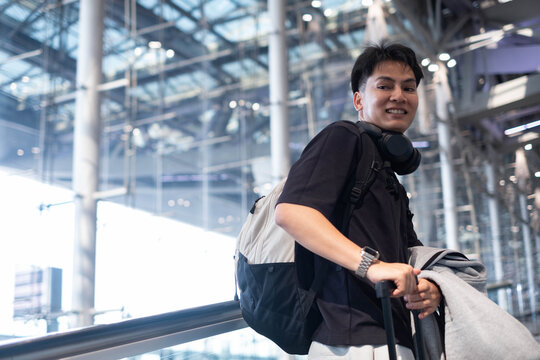 Happy young traveler walking through a modern airport with a backpack, headphones, and suitcase, smiling at the camera while preparing for a vacation, business trip, or international journey.