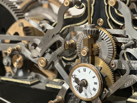 Intricate brass and steel clockwork gears surround a white clock face featuring traditional Roman numerals in a detailed close shot.