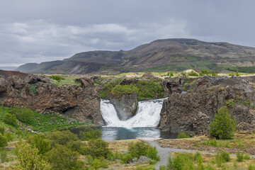 Fototapeta premium Hjalparfoss waterfall in the volcanic landscape of Thjorsardalur in wild Icelandic scenery. Gentle daylight preserves subtle earth and green tones. The shrubs define this vivid volcanic landmark.