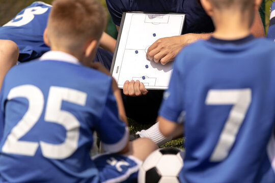 Youth Soccer Coach Pointing to Tactical Clipboard Diagram with Players Numbers 25 and 7 Listening - Football Team Strategic Meeting on Pitch