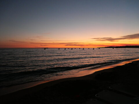 Coucher de soleil paisible sur l'oc&eacute;an avec des teintes orange et roses se refl&eacute;tant sur l'eau et de petits bateaux en silhouette &agrave; l'horizon.